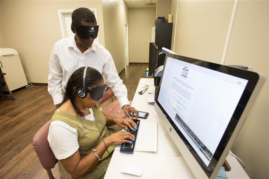 A student types at a computer with the assistance of the student standing behind her.